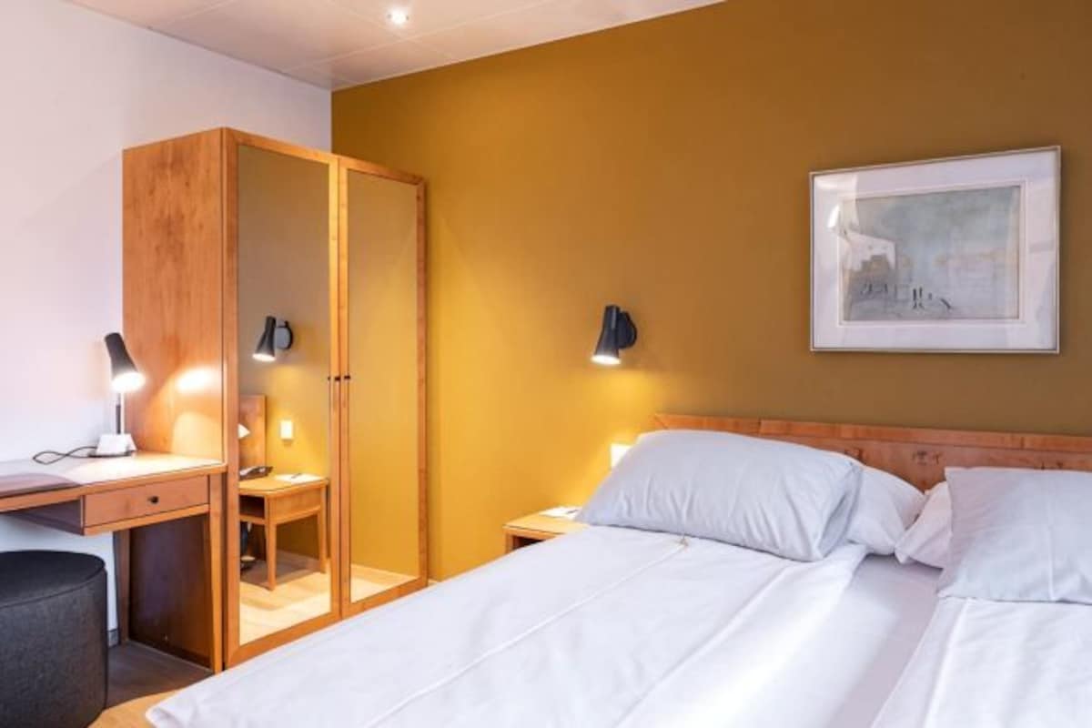 A hotel room with a bed, a desk, a wardrobe, and a nightstand with a lamp.