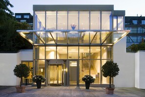 A modern hotel entrance with a glass facade, a central staircase, and two potted trees. - Engimatt City & Garden Hotel (Zürich)