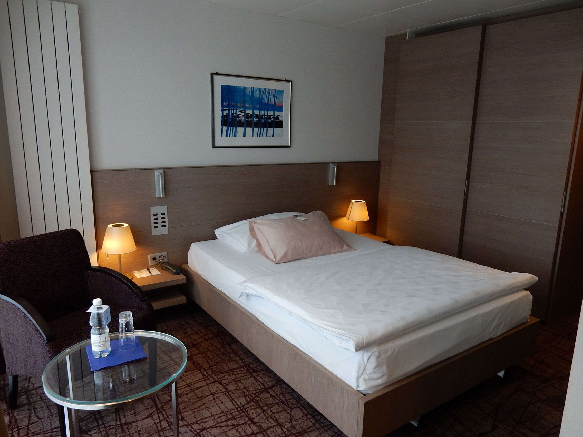 Ein Hotelzimmer mit einem Bett, Nachttischlampen, einem Sessel, einem Glastisch mit Wasserflaschen, einem gerahmten Bild an der Wand und einem Kleiderschrank.