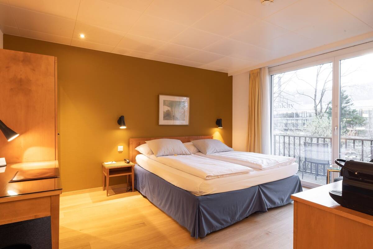 A hotel room with a bed, bedside tables, a desk, and a window with a view of the outside.