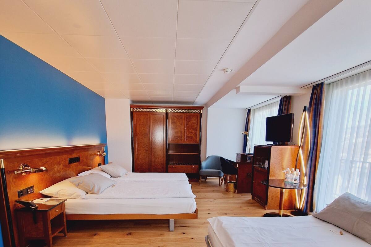A hotel room with two beds, a wooden floor, a wardrobe, a desk with a chair, and a television.