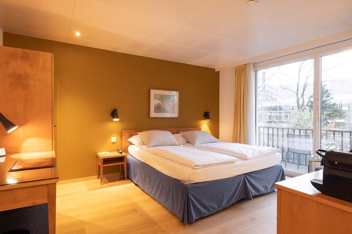 A hotel room with a bed, bedside table, lamp, and a view of the outside.