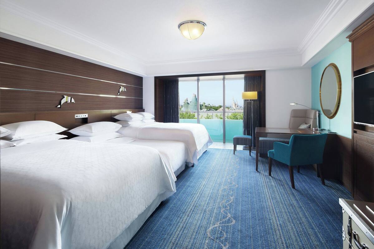 A hotel room with two beds, a desk, a chair, a TV, and a large window with a view.