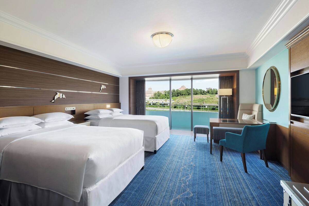 A hotel room with two beds, a desk, a chair, a TV, and a large window with a view of greenery and a building.