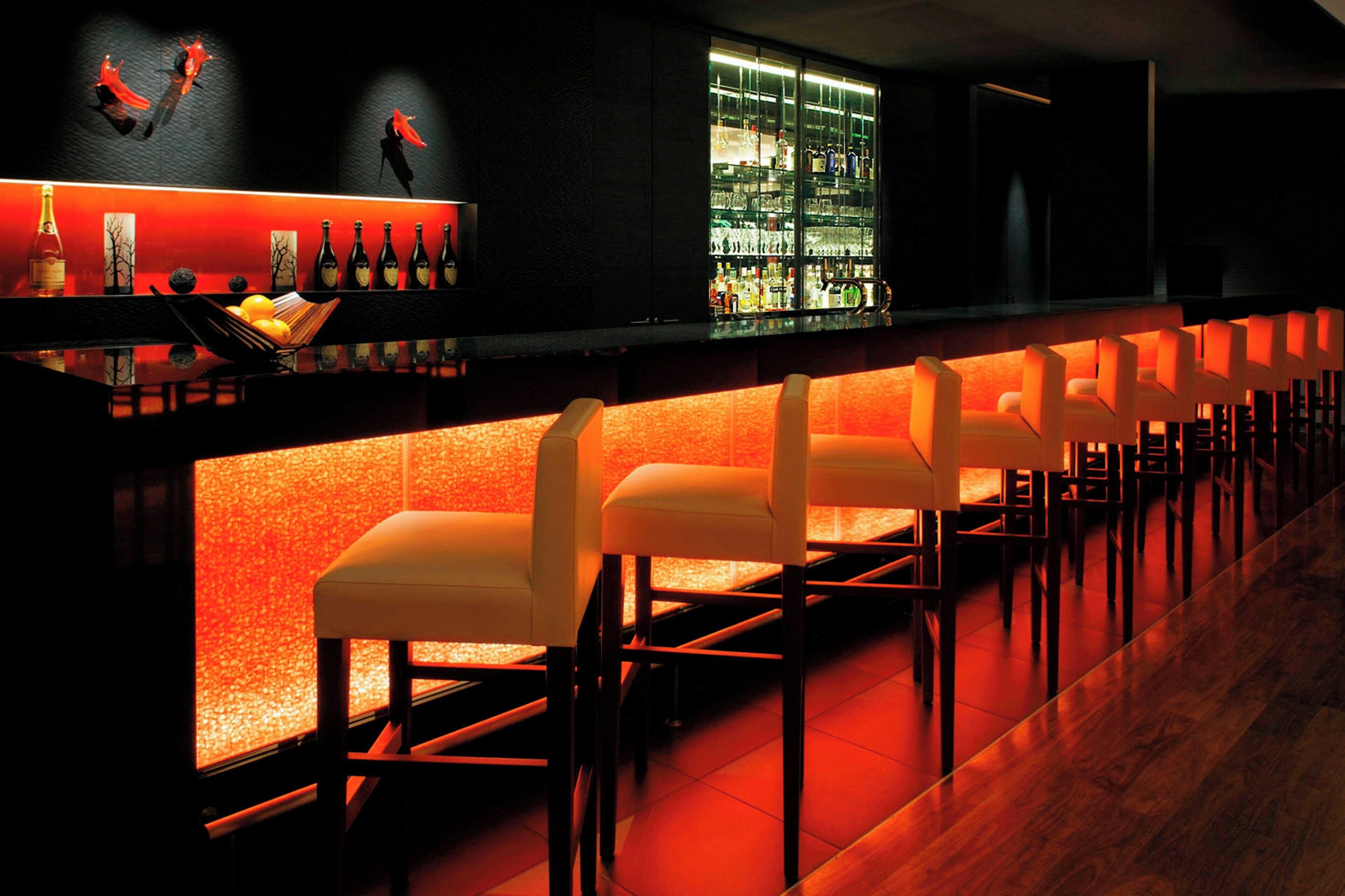 A bar area with a long counter, multiple seating options, and a well-stocked shelf with various bottles.