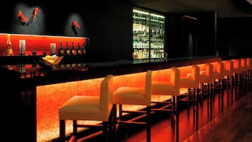 A bar area with a long counter, multiple seating options, and a well-stocked shelf with various bottles.