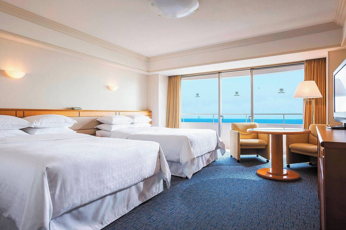 A hotel room with two beds, a small table, a chair, and a view of the ocean.