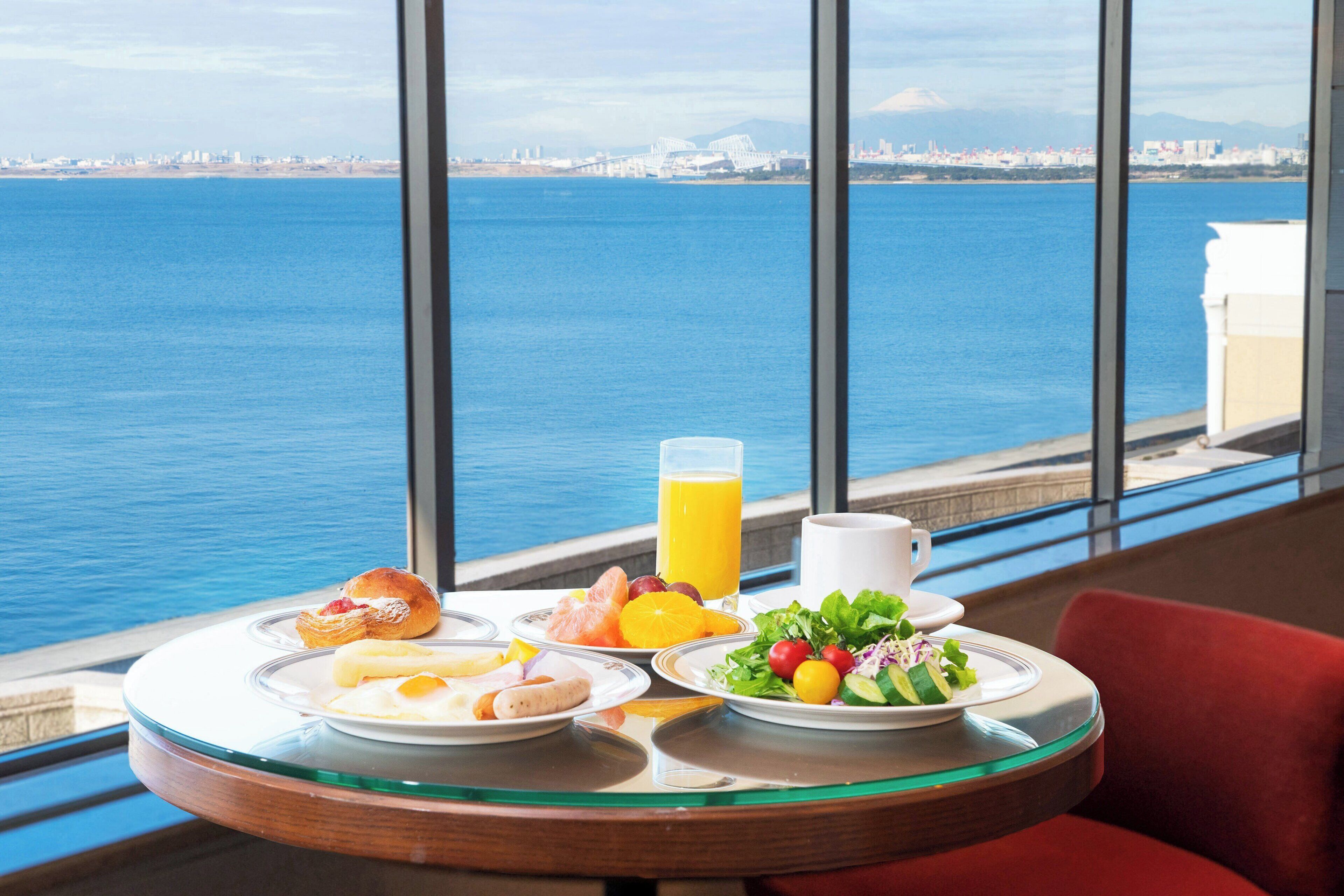 A breakfast table with plates of food, a glass of orange juice, and a view of the sea and cityscape.