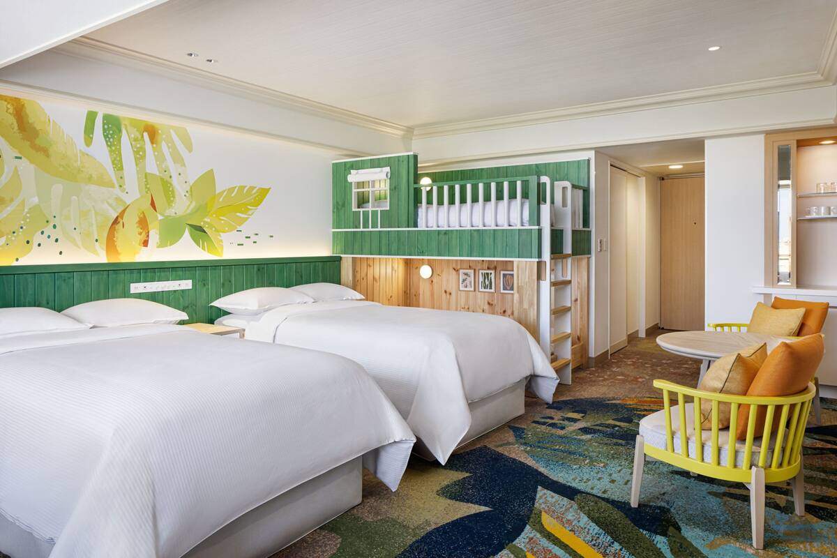 A hotel room with two beds, a green wall with a leaf mural, a wooden bunk bed, a yellow chair, and a round table.