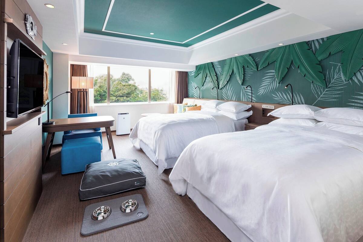 A hotel room with two beds, a TV, a desk with a chair, a window with a view, and a ceiling with a leaf pattern.