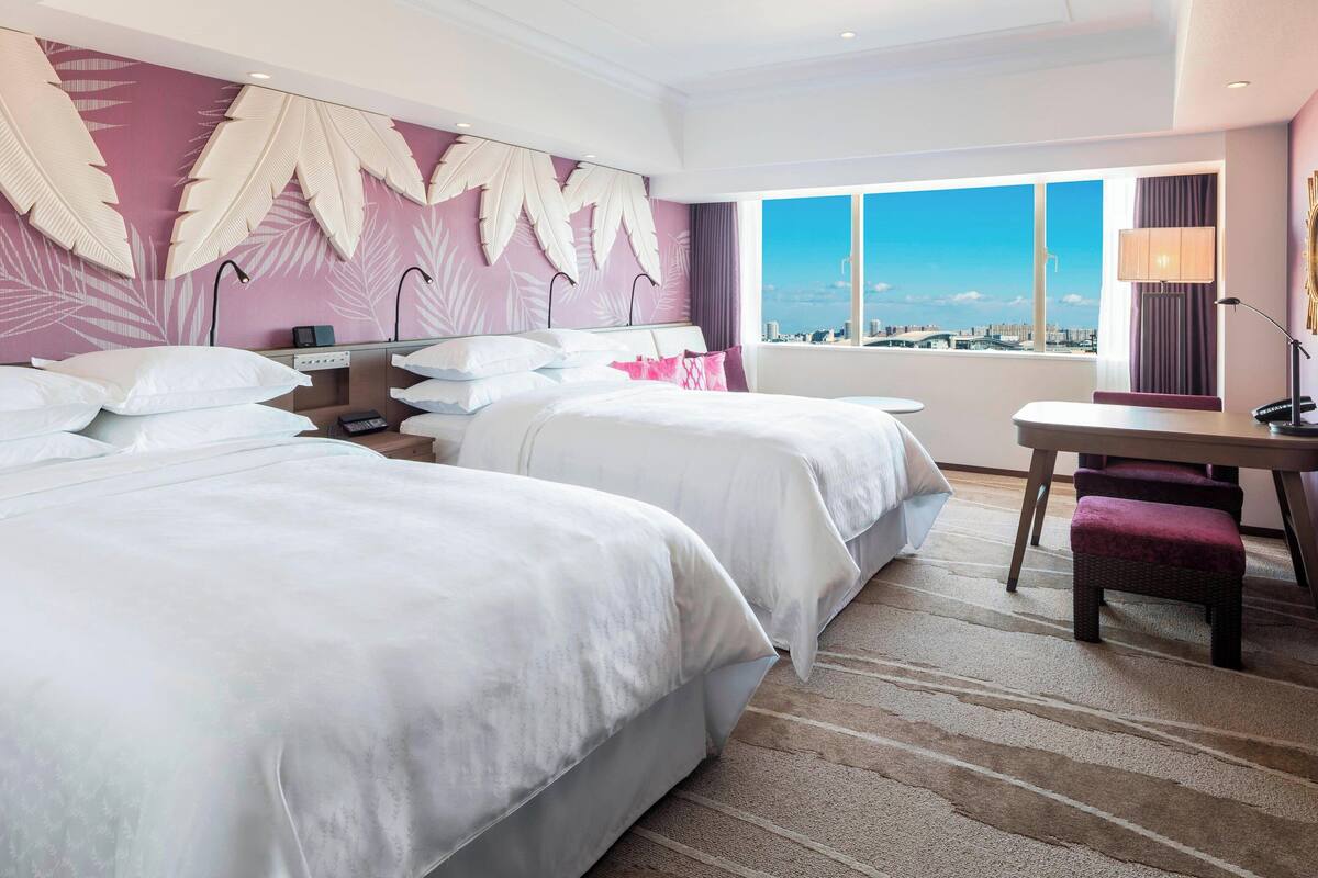 A hotel room with two beds, a desk, and a large window offering a city view.