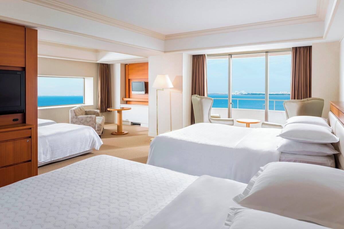 A hotel room with two beds, a TV, a desk, and a view of the sea.