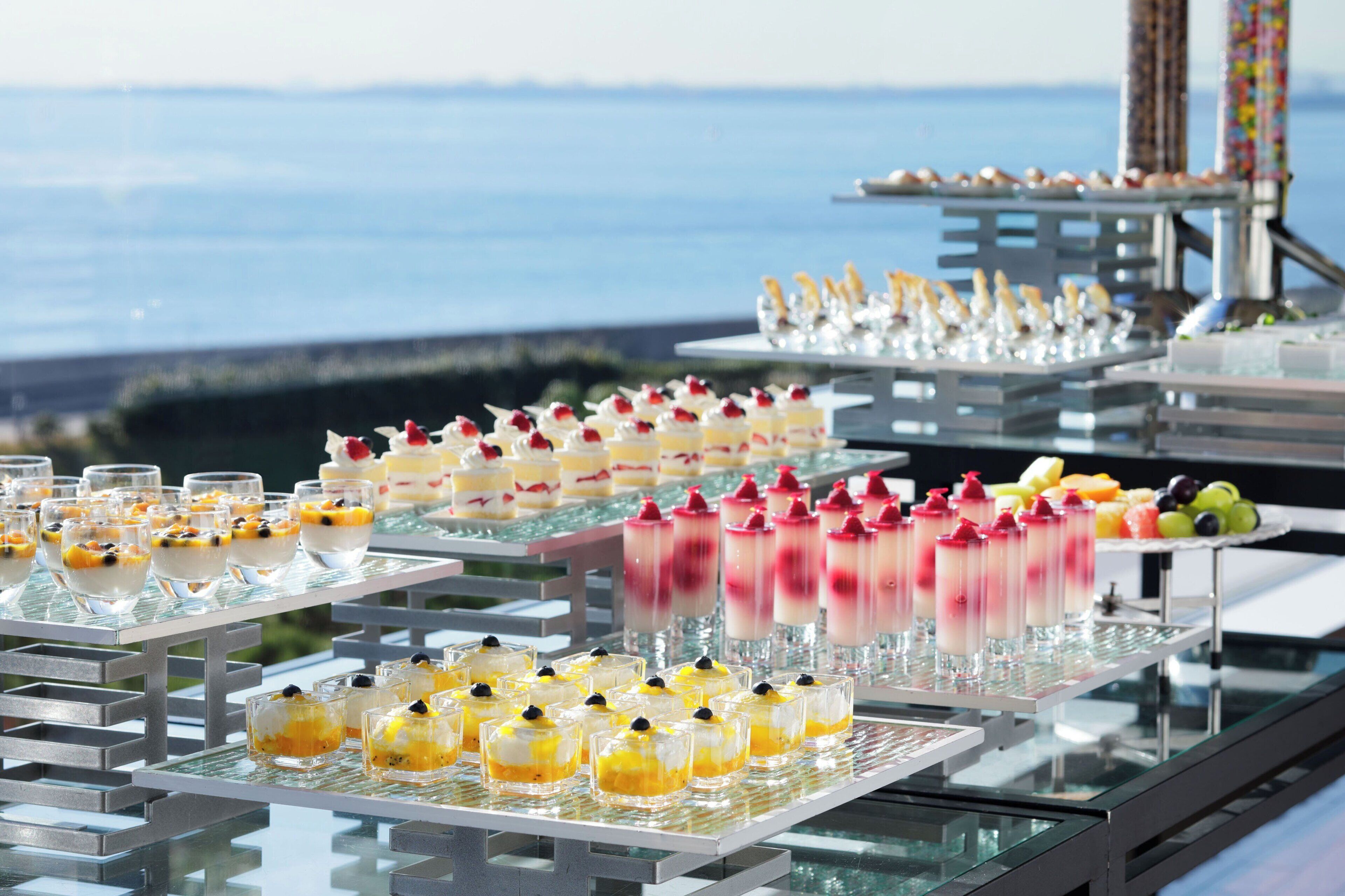 Un bufé con diversos postres y frutas, con el cielo despejado y una masa de agua de fondo.