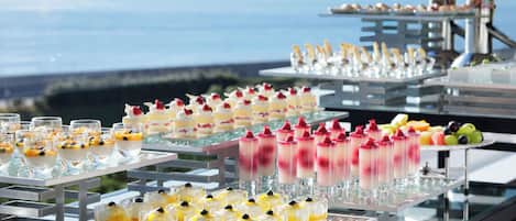 A buffet with various desserts and fruits, set against a backdrop of a clear sky and a body of water.