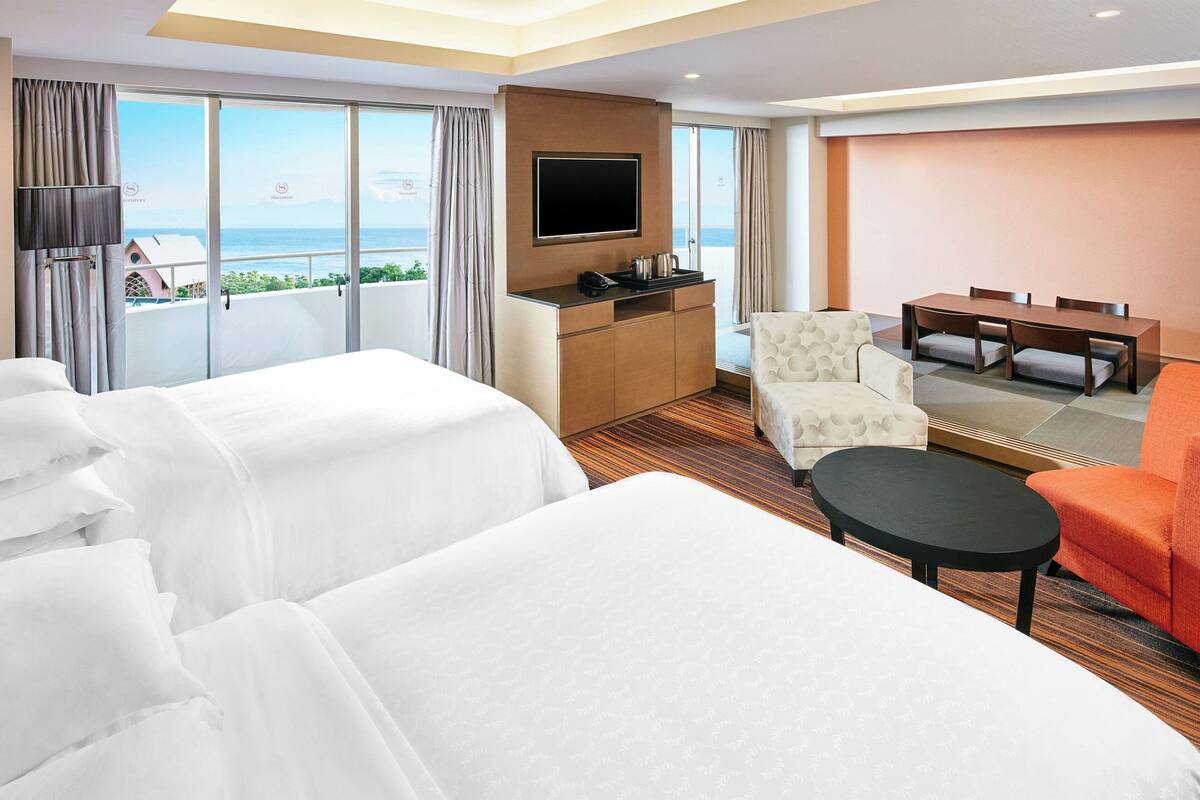 A hotel room with two beds, a TV, a small dining table, and a view of the sea.