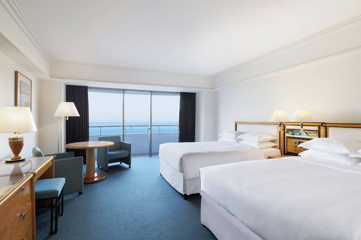 A hotel room with two beds, a desk with a lamp, a chair, a small table, and a view of the ocean through large windows.