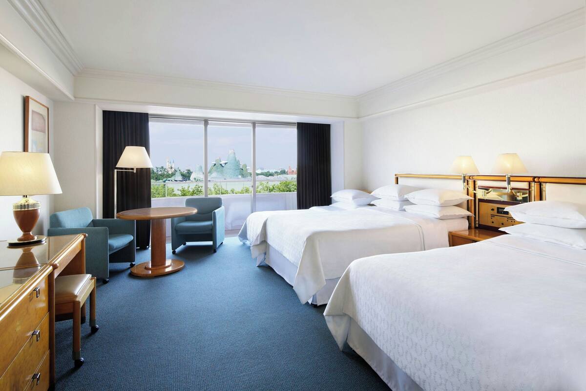 A hotel room with two beds, a desk, a chair, and a view of a park and buildings.