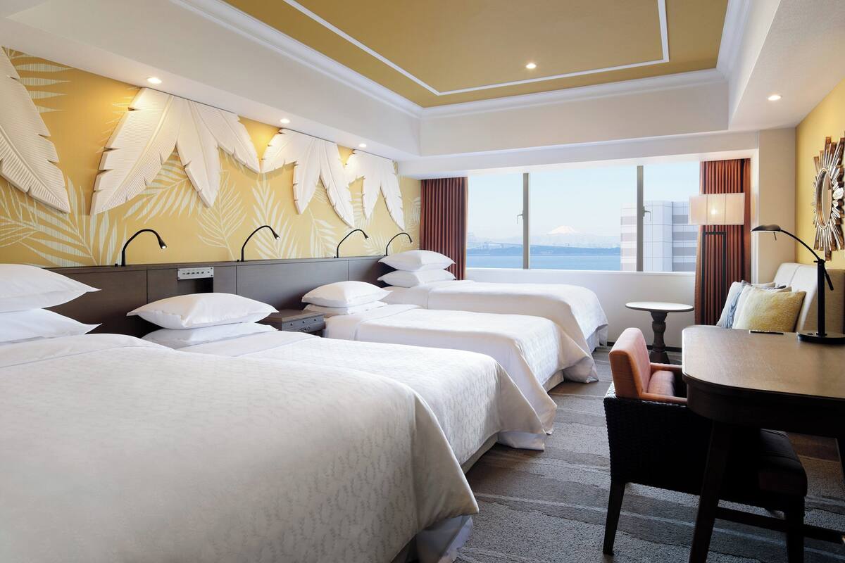 A hotel room with three beds, a dining table, and a view of the city.
