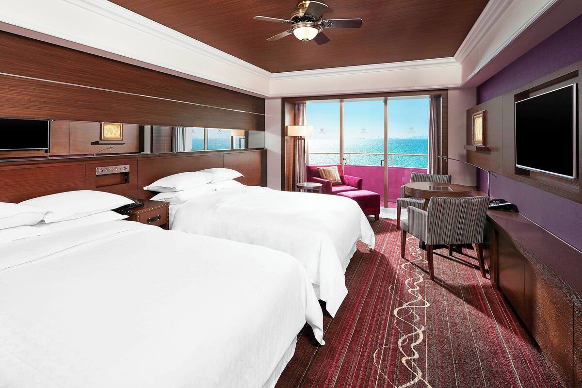 A hotel room with two beds, a desk, a chair, a TV, and a large window with a view of the sea.