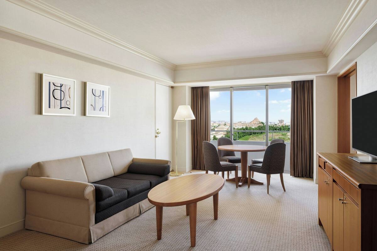A hotel room with a sofa, a dining table with chairs, a TV, and a large window with a view.