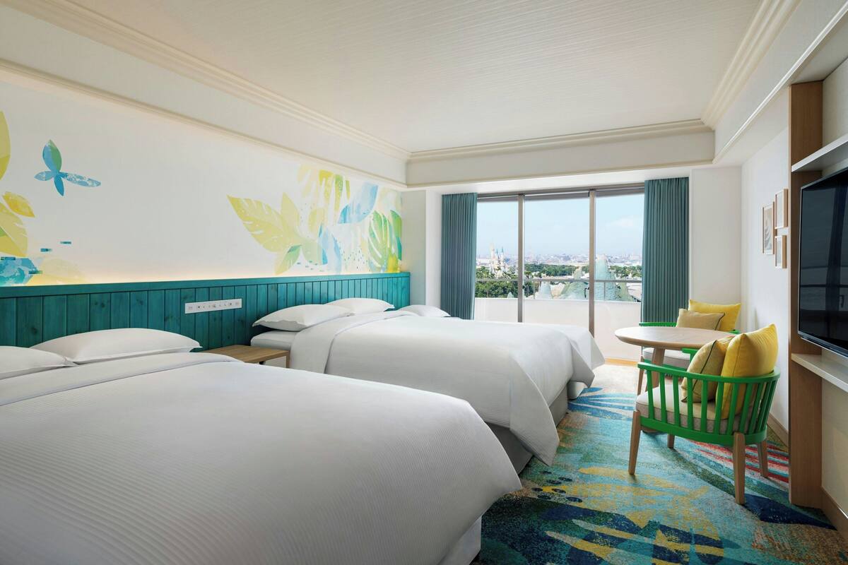 A hotel room with two beds, a small table with chairs, a TV, and a balcony with a view of the city.
