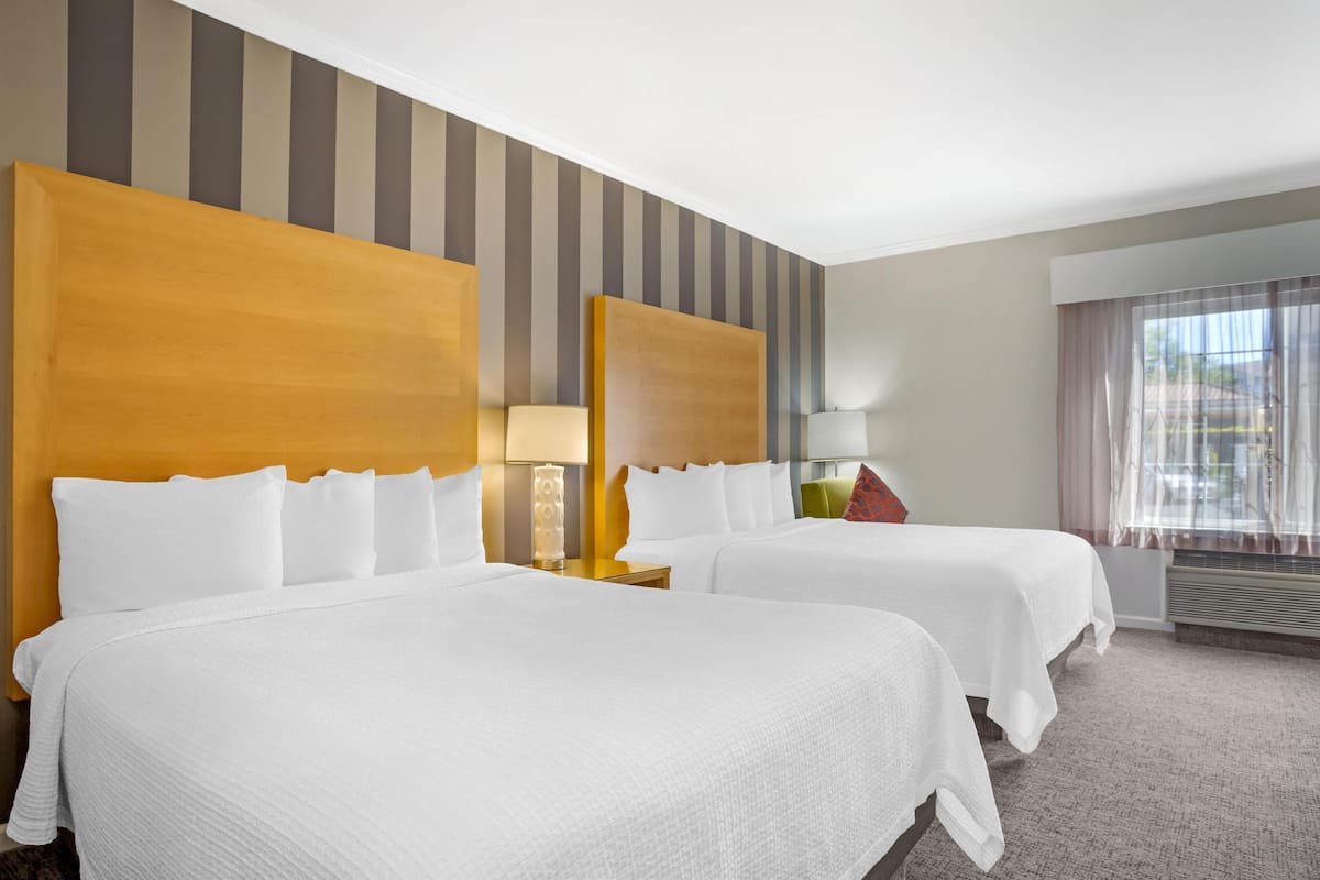 Une chambre d’hôtel avec deux lits, un papier peint à rayures, une fenêtre avec des rideaux et une lampe sur la table de chevet.