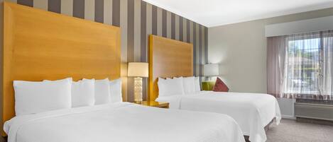 A hotel room with two beds, striped wallpaper, a window with curtains, and a lamp on the nightstand.