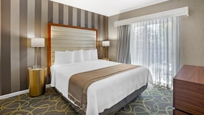 Une chambre d’hôtel avec un grand lit, deux lampes de chevet, une tête de lit en bois, une fenêtre avec des rideaux légers et une moquette à motifs.