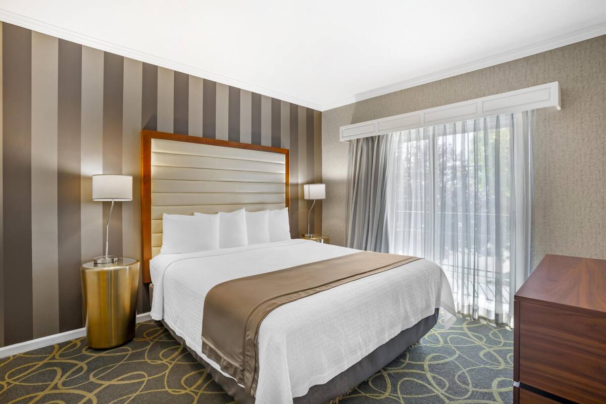 Une chambre d’hôtel avec un grand lit, deux lampes de chevet, une tête de lit en bois, une fenêtre avec des rideaux légers et une moquette à motifs.