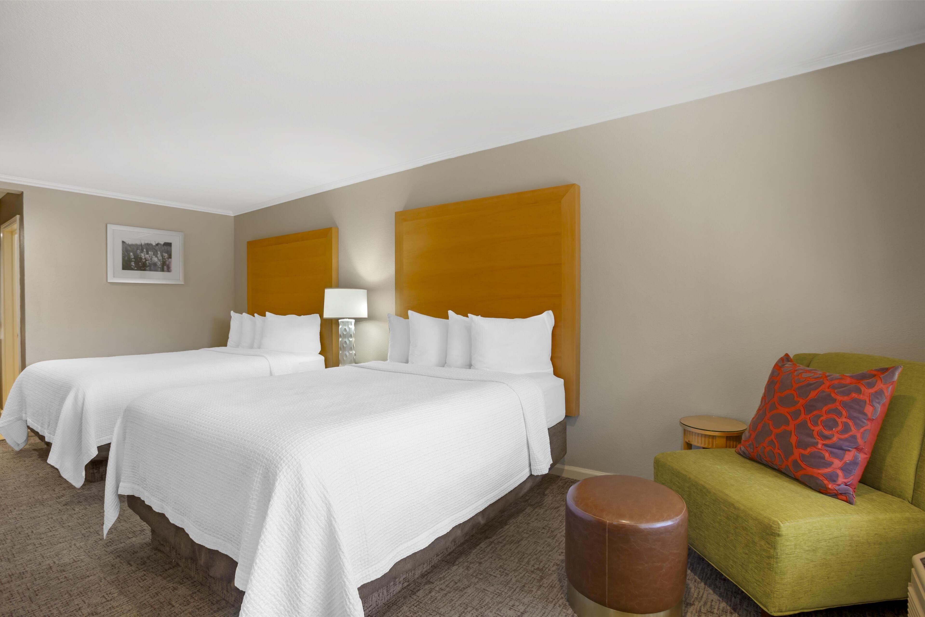 A hotel room with two beds, a green armchair, a small round table, and a lamp.