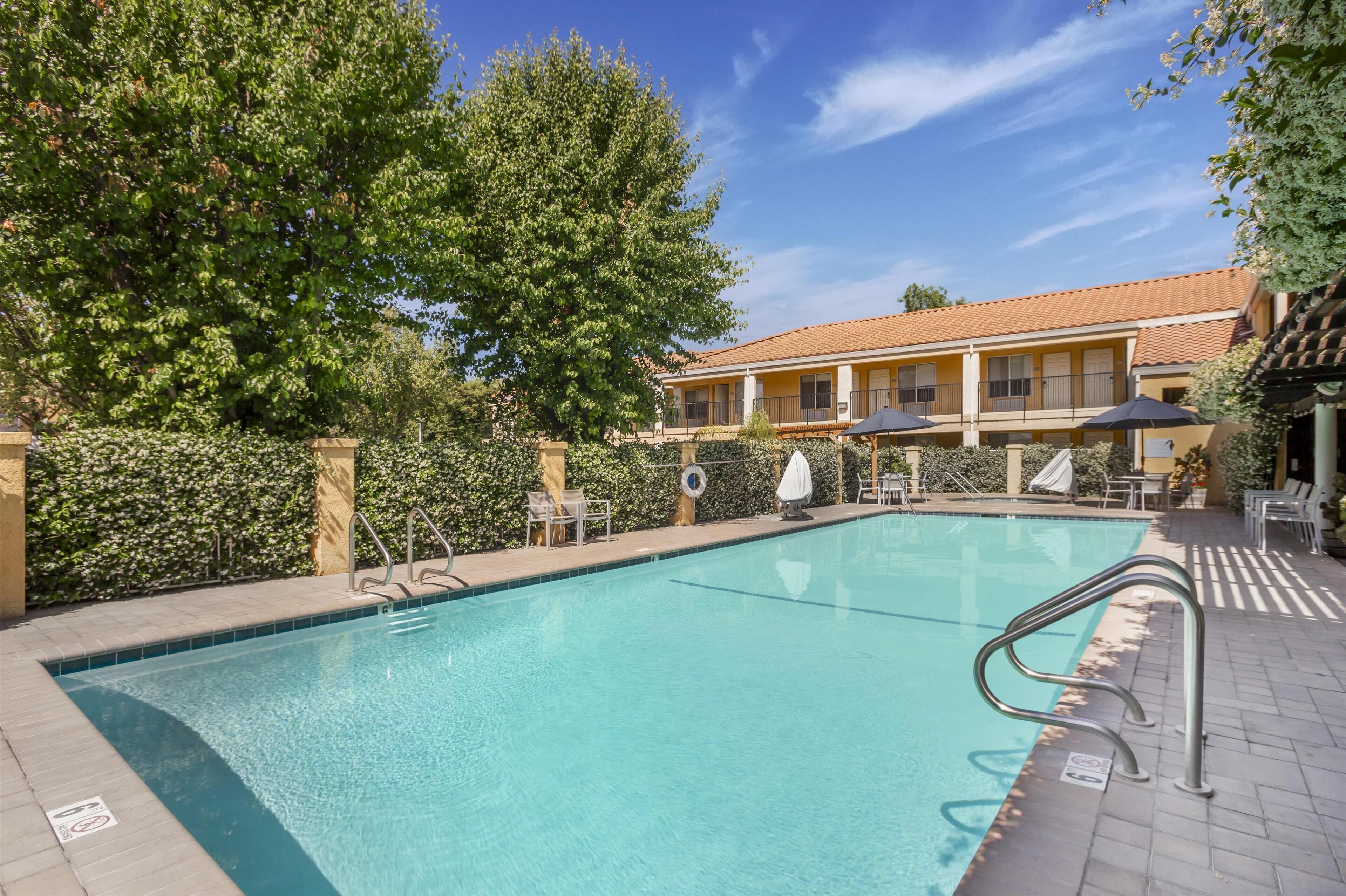 A swimming pool with a tiled deck, surrounded by a hedge and a building with a tiled roof.