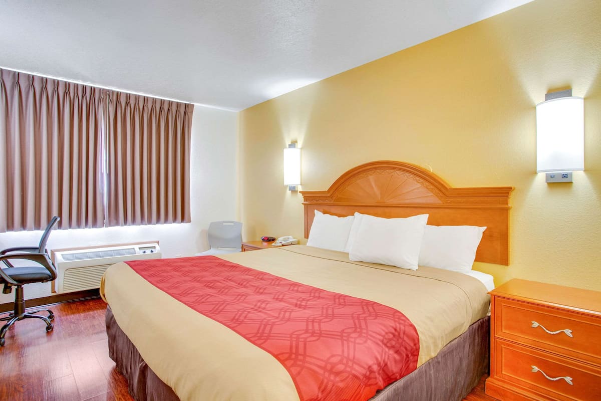 A hotel room with a large bed, a wooden nightstand, a desk with a chair, and a window with curtains.