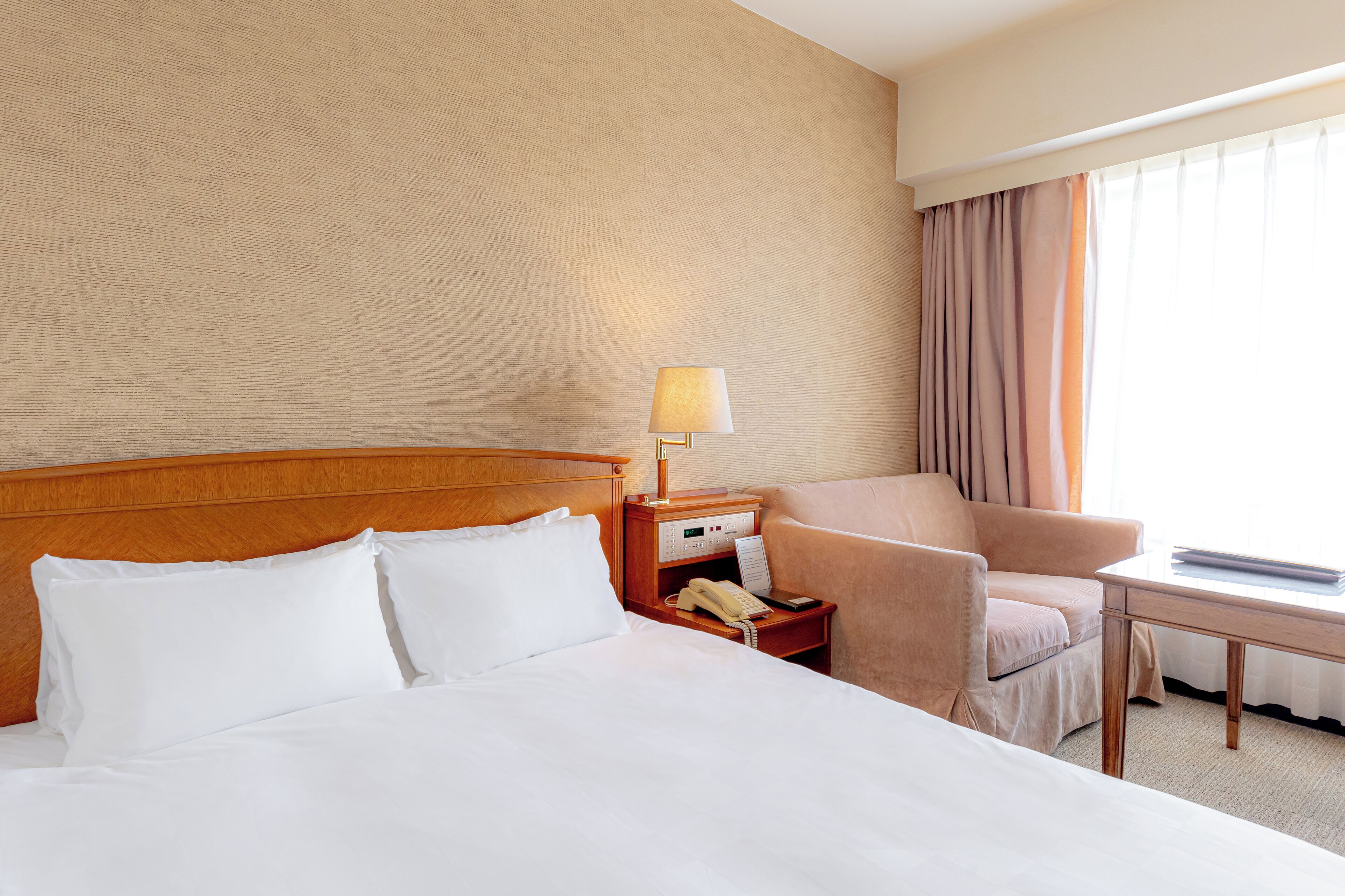 Quarto de hotel com uma cama grande, cabeceira de madeira, mesa de cabeceira com abajur, telefone e um espaço de estar com uma cadeira e uma mesa.