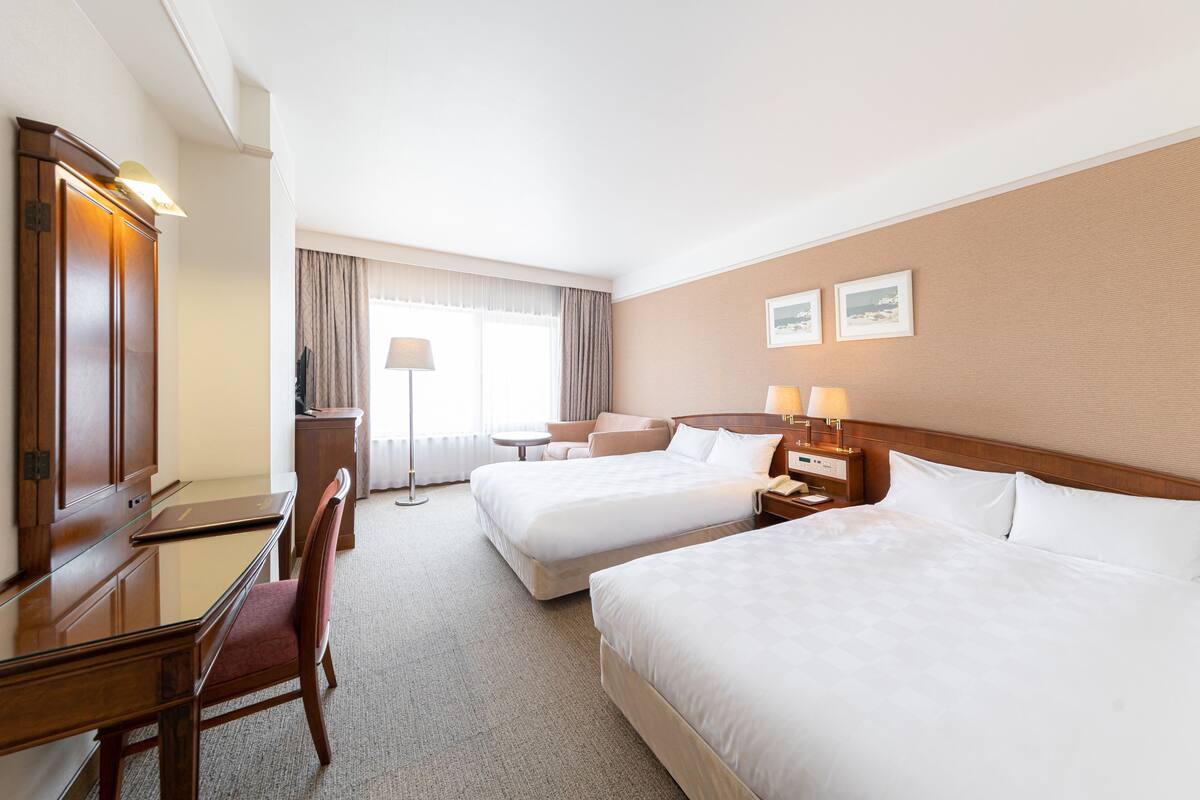 A hotel room with two beds, a desk, a chair, a lamp, and a window with curtains.