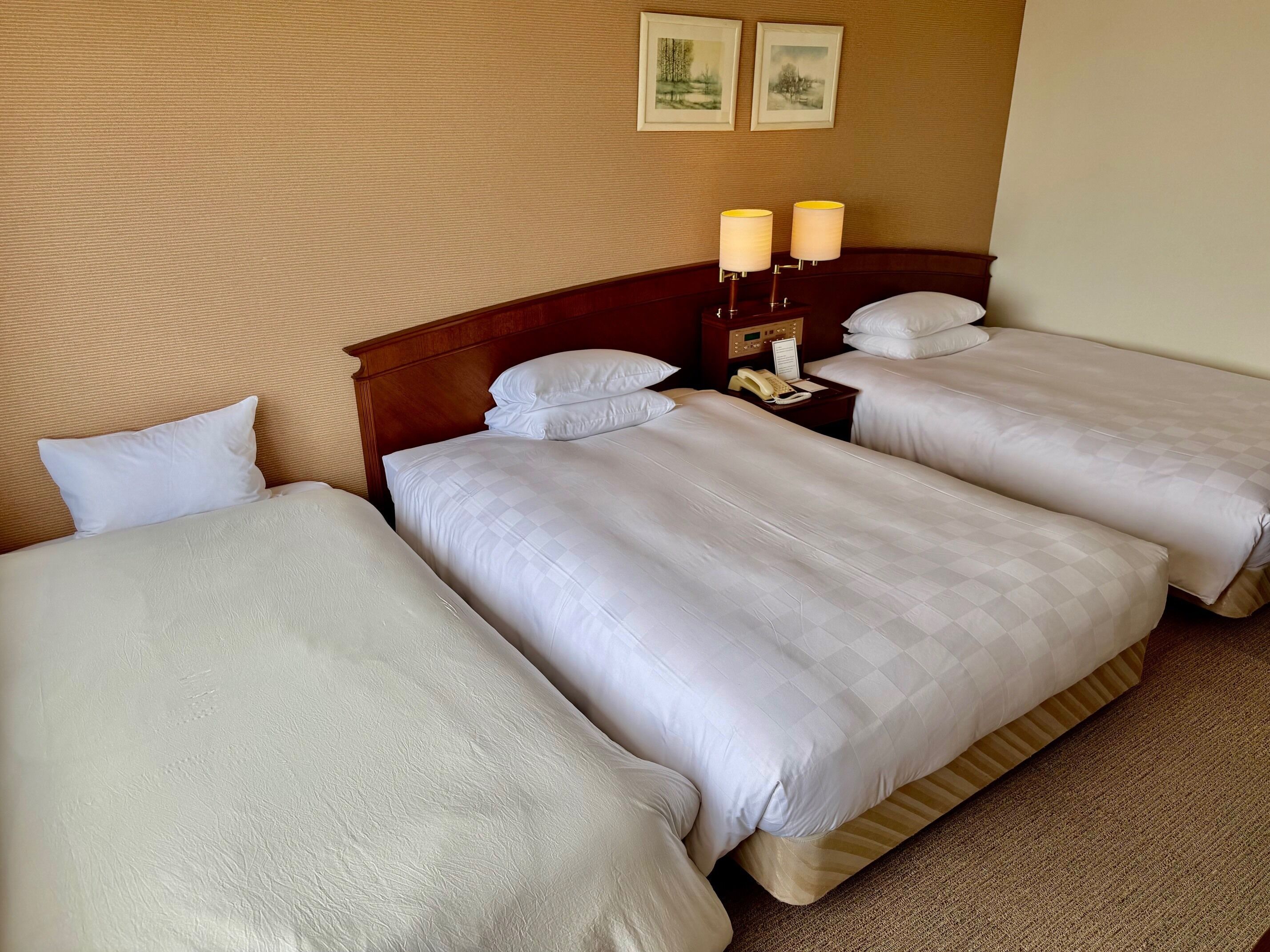 A hotel room with three beds, a wooden headboard, a nightstand with lamps, and two framed pictures on the wall.