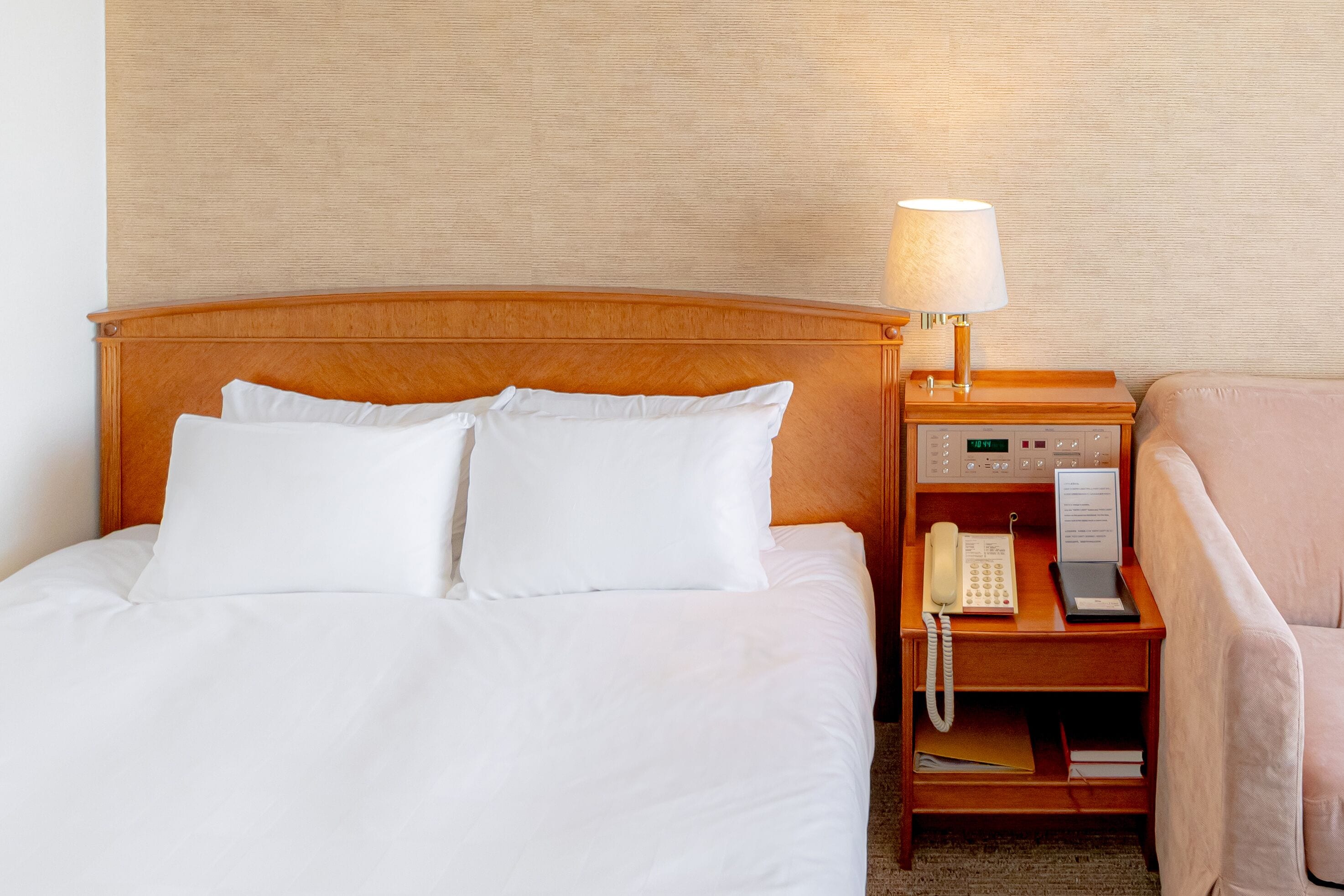 A hotel room with a wooden headboard bed, white bedding, a bedside table with a lamp, a telephone, and a chair.