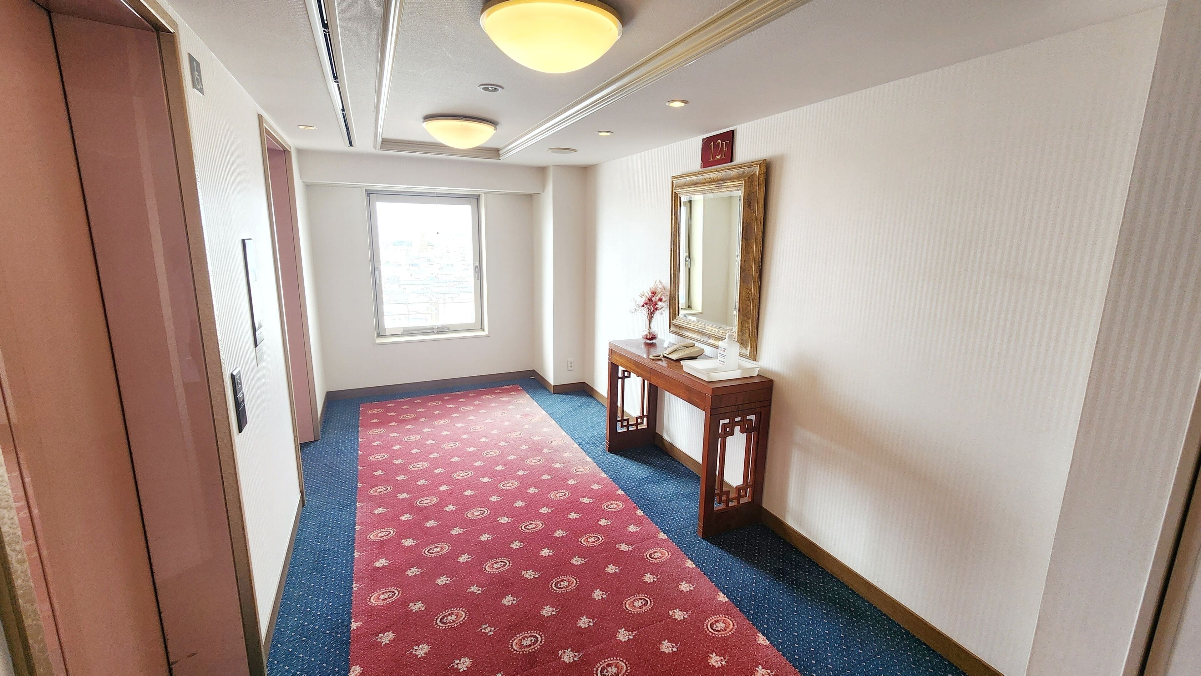 A hotel hallway with a red carpet, a wooden console table with a mirror, and a window providing natural light.