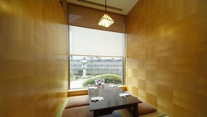 Una habitación japonesa tradicional con suelo de tatami, una mesa baja con cojines y vistas a un jardín por una ventana grande.