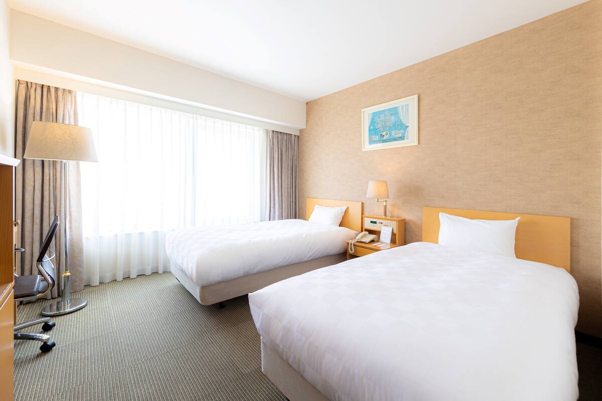 A hotel room with two beds, a desk with a lamp, a chair, and a window with curtains.