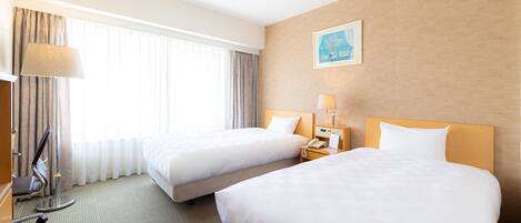A hotel room with two beds, a desk with a lamp, a chair, and a window with curtains.