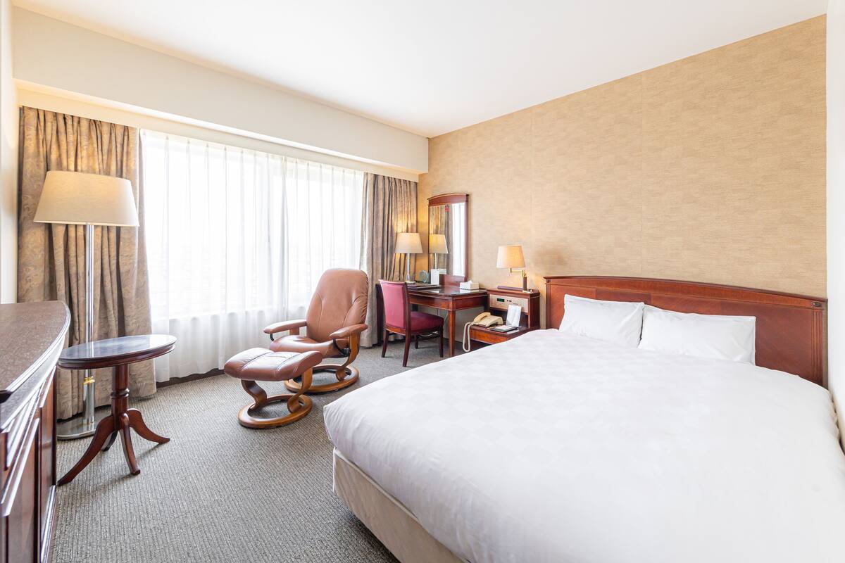 A hotel room with a large bed, a desk with a telephone, a chair, a lamp, and a window with curtains.