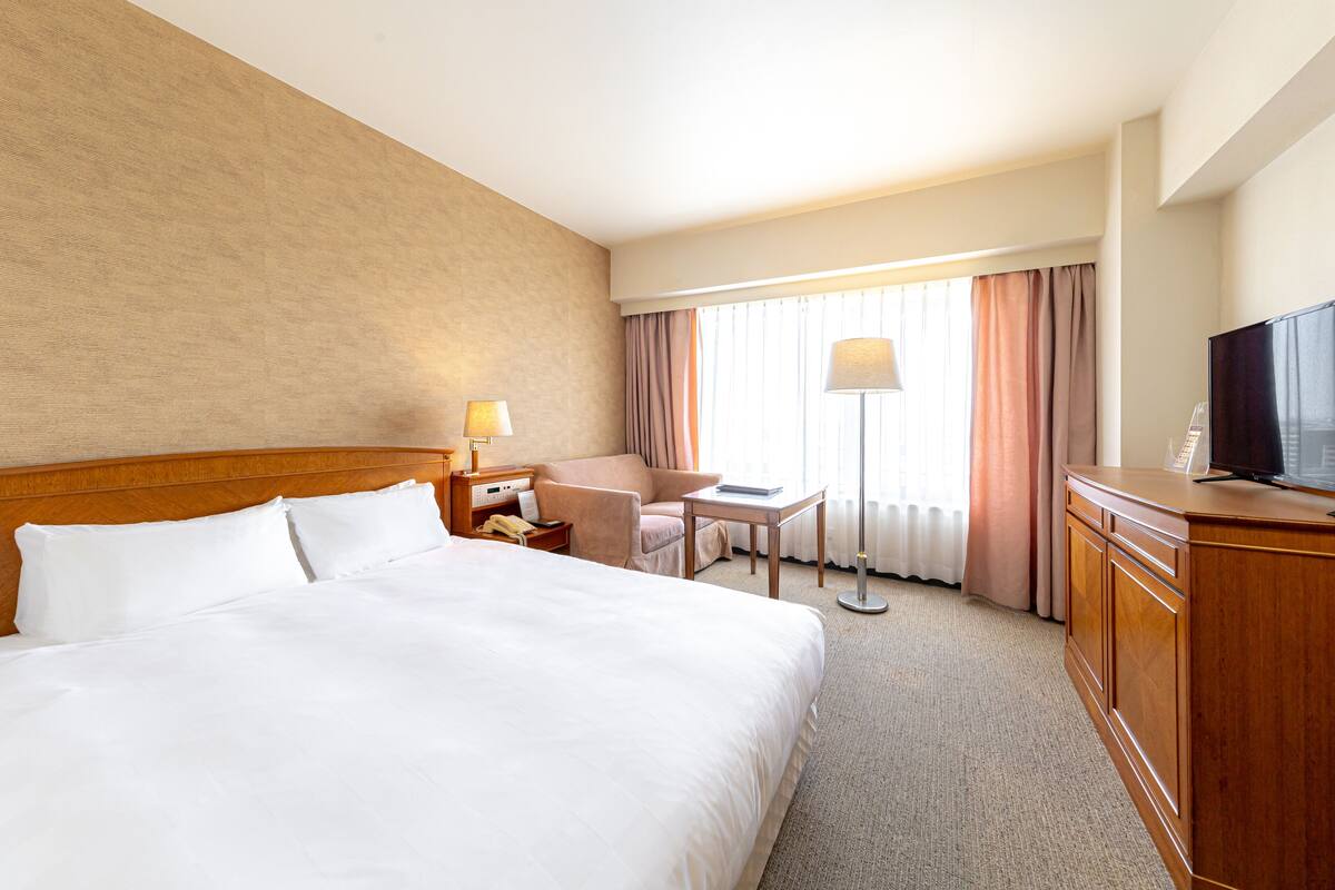 A hotel room with a large bed, a desk, a chair, a TV, and a window with curtains.