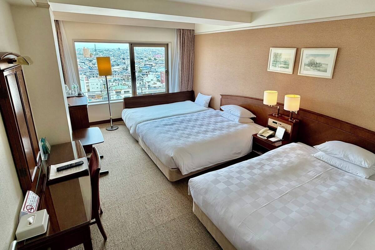 A hotel room with two beds, a desk, a chair, a lamp, a phone, and a window with a city view.