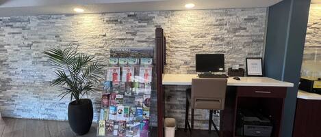 Zona de recepción con pared de piedra, escritorio con ordenador, silla, planta en maceta y un mueble para revistas.