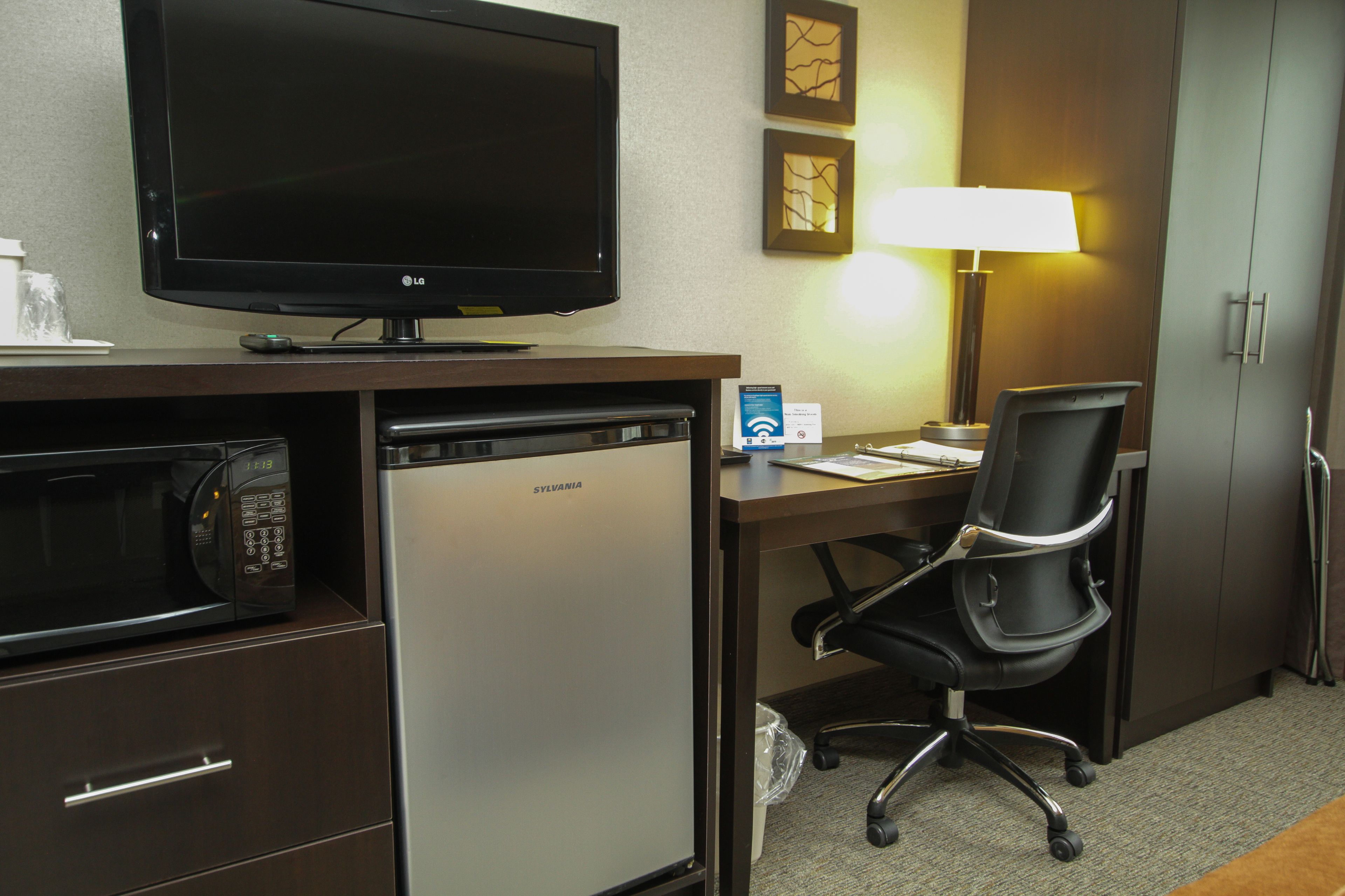 Habitación de hotel con televisión de pantalla plana, microondas, refrigerador pequeño, escritorio con silla, lámpara y armario.