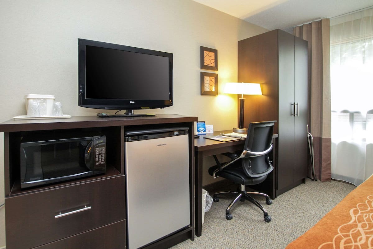 A hotel room with a flat-screen TV, a microwave, a mini-fridge, a desk with a chair, a lamp, and a bed with a patterned bedspread.