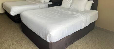 A hotel room with two single beds, a dark headboard, a nightstand with a lamp, and two framed pictures on the wall.