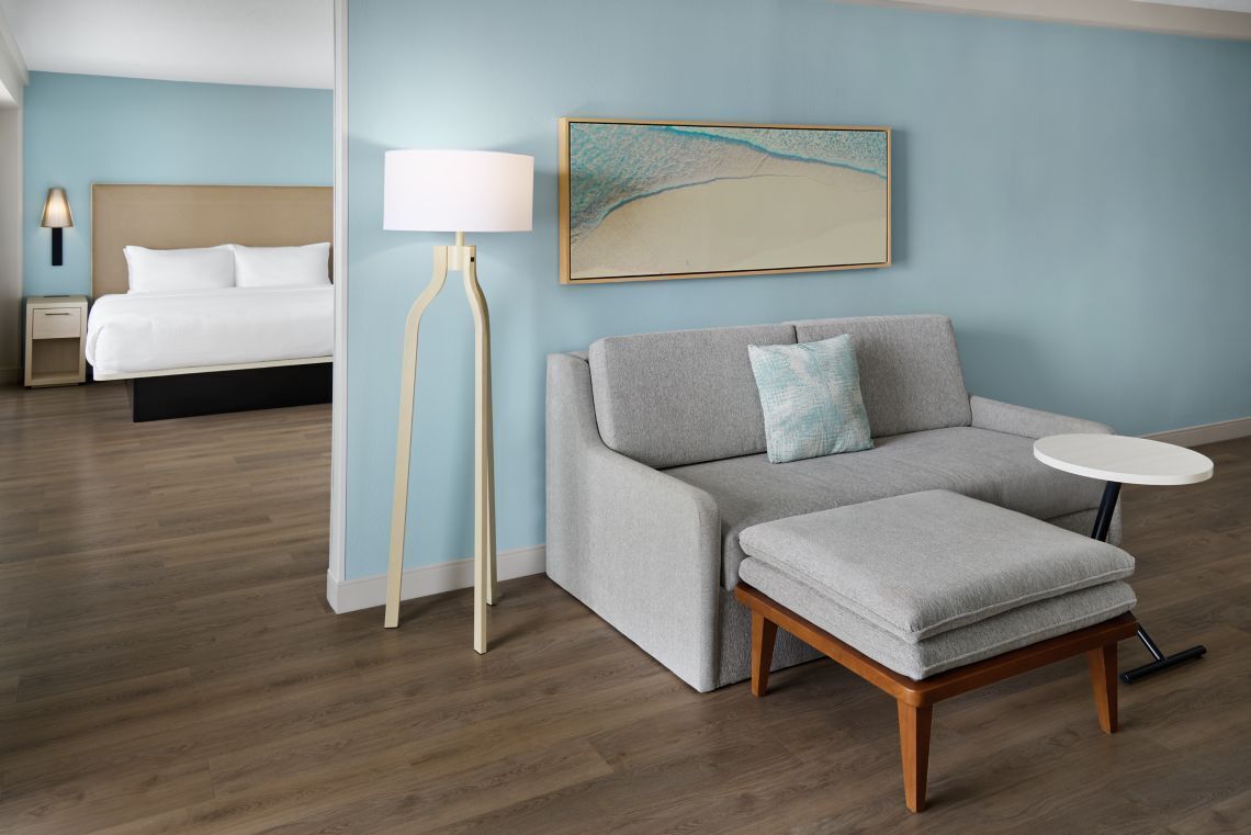 Ein Hotelzimmer mit einem Bett, einem grauen Sofa, einem kleinen runden Tisch, einer Stehlampe und einem Wandbild mit Strandmotiv.