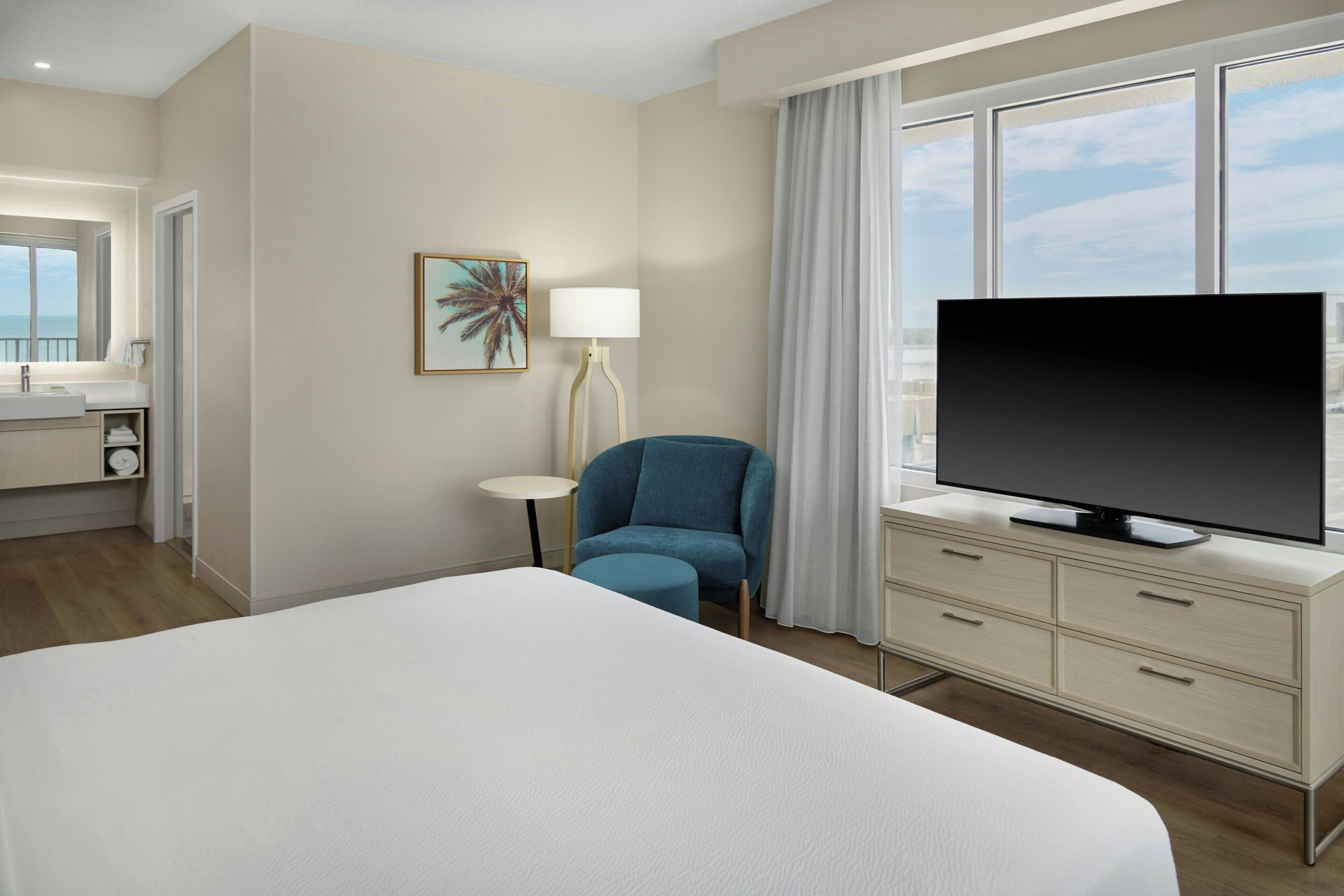 Habitación de hotel con cama, televisor en una cómoda, una silla, una mesita y ventana con cortinas.