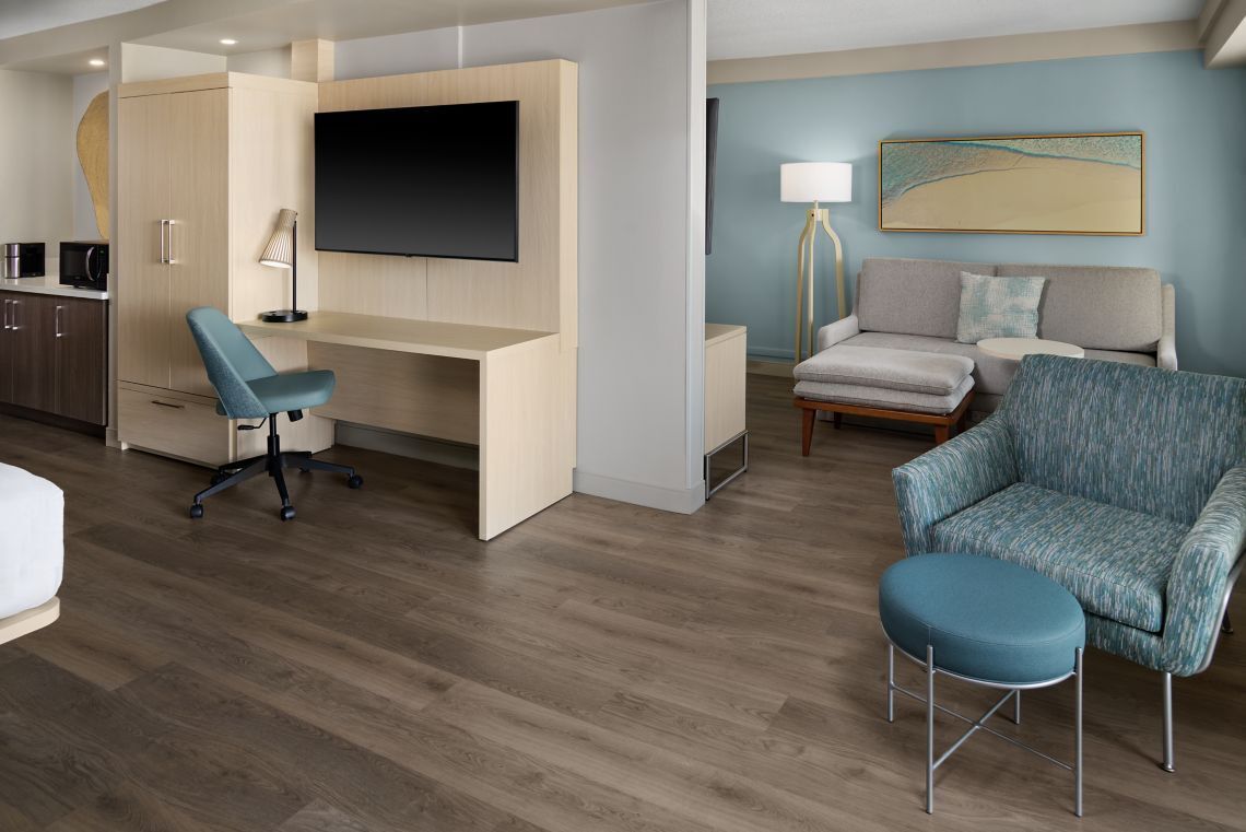 A modern hotel room with a wooden floor, a desk with a chair, a sofa, a small ottoman, a TV mounted on the wall, and a lamp.